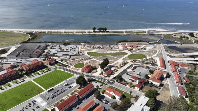 A sweeping aerial daylight view of Presidio Tunnel Tops park featuring the uplands visitor center, the Crissy Field Marsh, and the San Francisco Bay with sail boats on the water