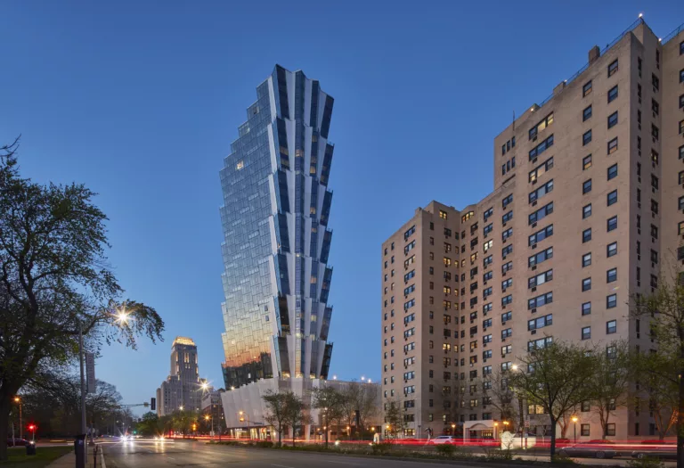 Exterior twilight view of the flare-tiered, 36-story One Hundred Above the Park’s glass facade with a five-story podium and roadway traffic in the foreground