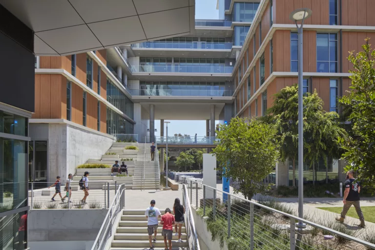 Exterior daylight view of students walking through the plaza and up staircases at the University of California San Diego’s North Torrey Pines, surrounded by a mid-rise building with a glass-and-wood facade