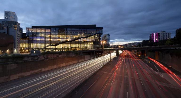 Exterior twilight view of the Seattle Convention Center Addition illuminated from within, featuring the Pine Street overpass and streaming headlights and taillights of vehicle traffic along Interstate 5