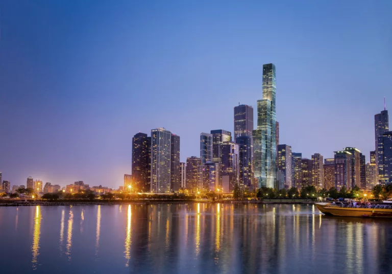 Exterior twilight view of the 12,000-foot-tall, 100-story St. Regis Chicago surrounded by the city’s illuminated skyline and, in the foreground, a passenger boat on Lake Michigan