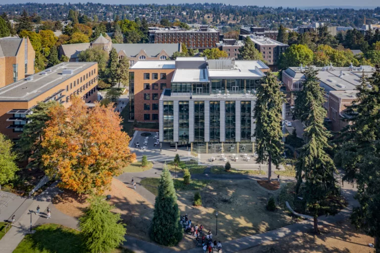Aerial daylight view of the University of Washington Foster School of Business’ Founders Hall, a five-story structure surrounded by trees, other academic buildings, and students walking along campus trails
