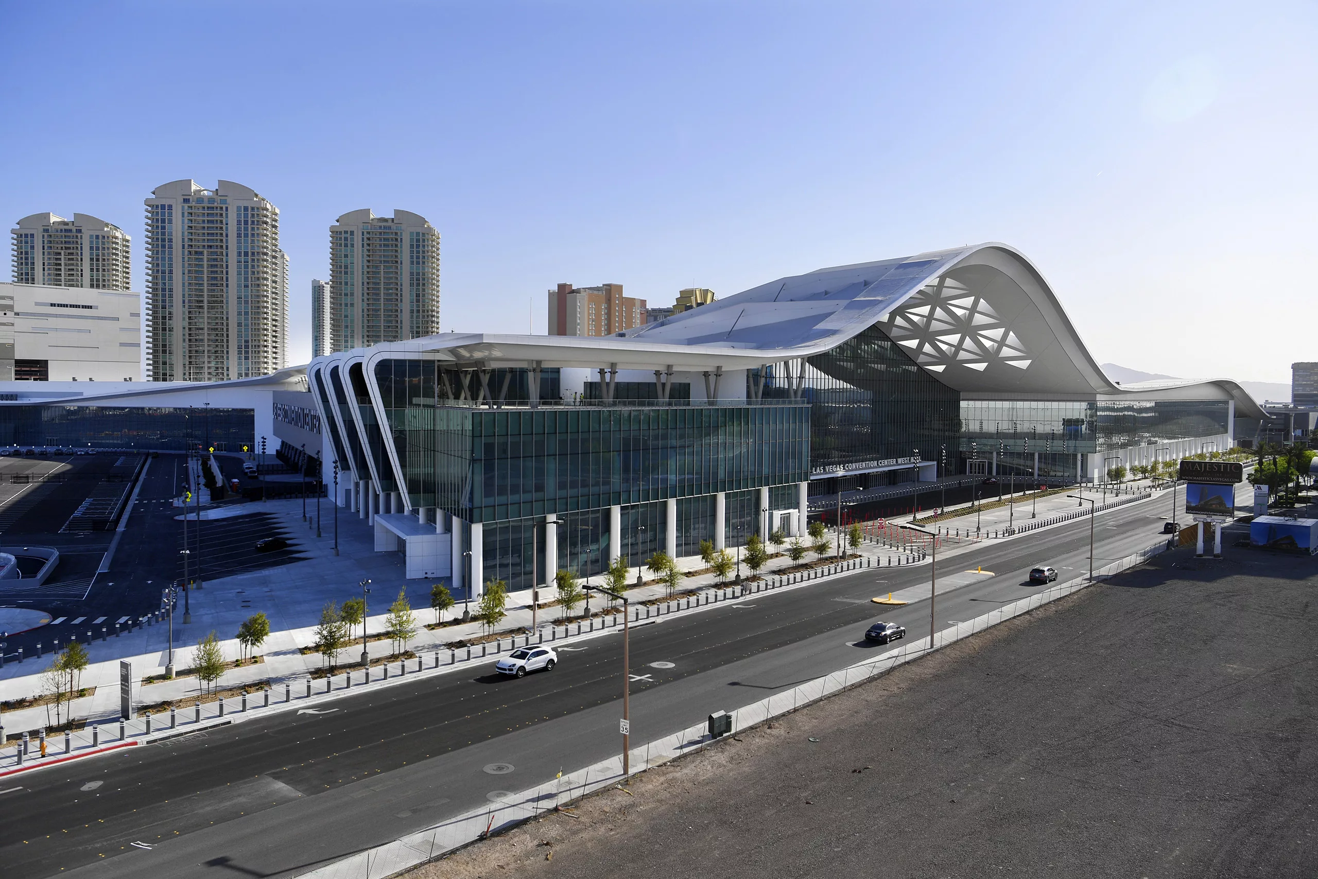 Aerial daylight view of the Las Vegas Convention Center Expansion's West Hall featuring an undulating roof, entry plaza, tree-lined sidewalk, and four-lane roadway with several high-rise buildings in the distance