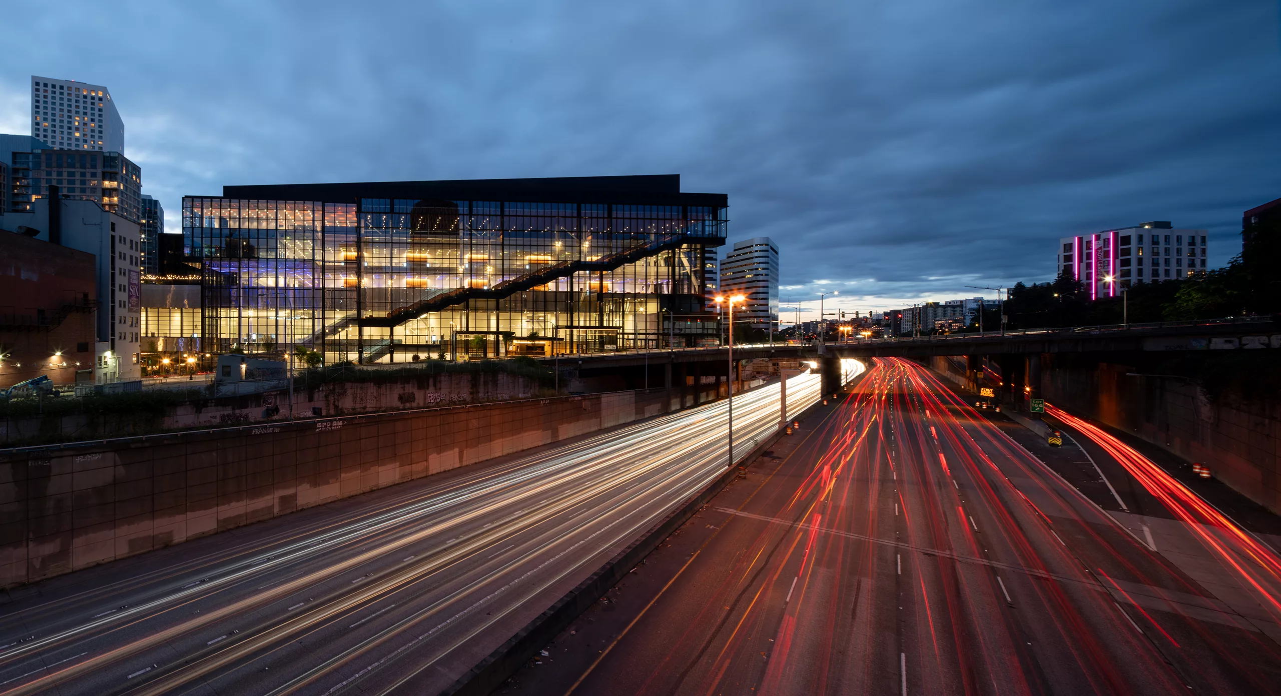 Exterior twilight view of the Seattle Convention Center Addition illuminated from within, featuring the Pine Street overpass and streaming headlights and taillights of vehicle traffic along Interstate 5