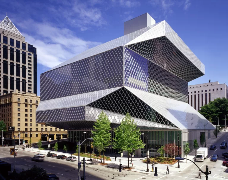 Slightly elevated daylight view of the west entry to the angular, steel-and-glass Seattle Central Library with surrounding streets, landscaping, and downtown buildings