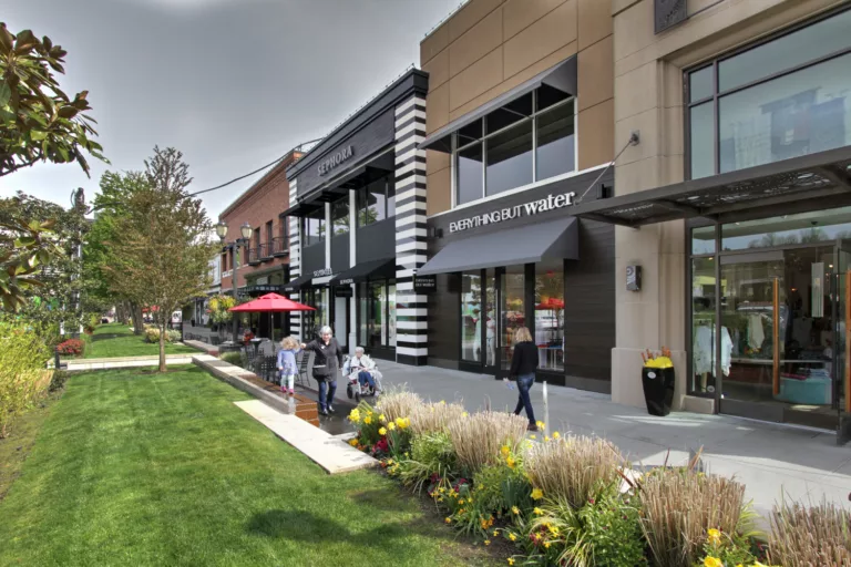 Exterior daylight view of University Village retail shops adjacent to a central plaza featuring outdoor seating, a water fountain, an expansive lawn, natural landscaping, and various shoppers