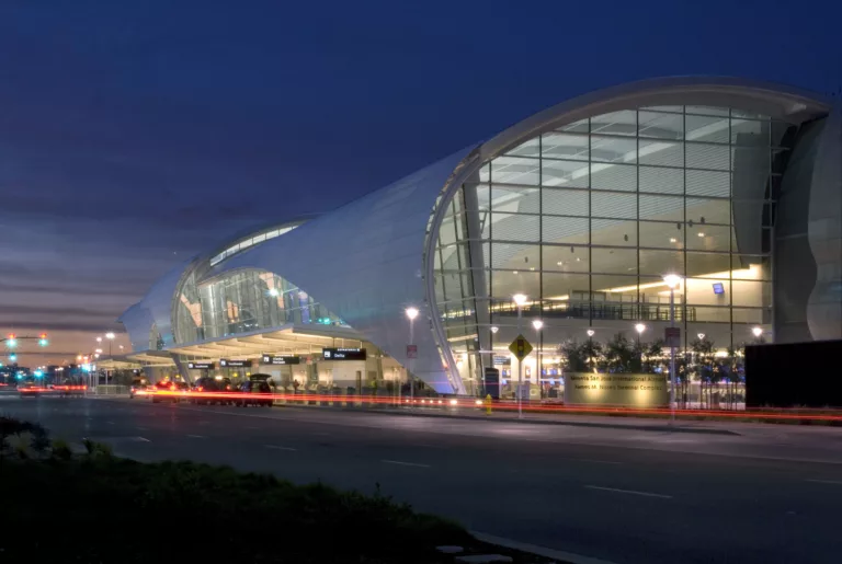 Exterior twilight view of Mineta International Airport's Terminal B featuring a towering glass facade partially wrapped in curved metal, with ticketing counters and security lines illuminated within