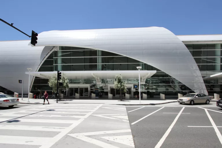 Exterior view of Mineta International Airport's Terminal B in daylight featuring a glass facade partially wrapped in curved metal and several vehicles parked curbside
