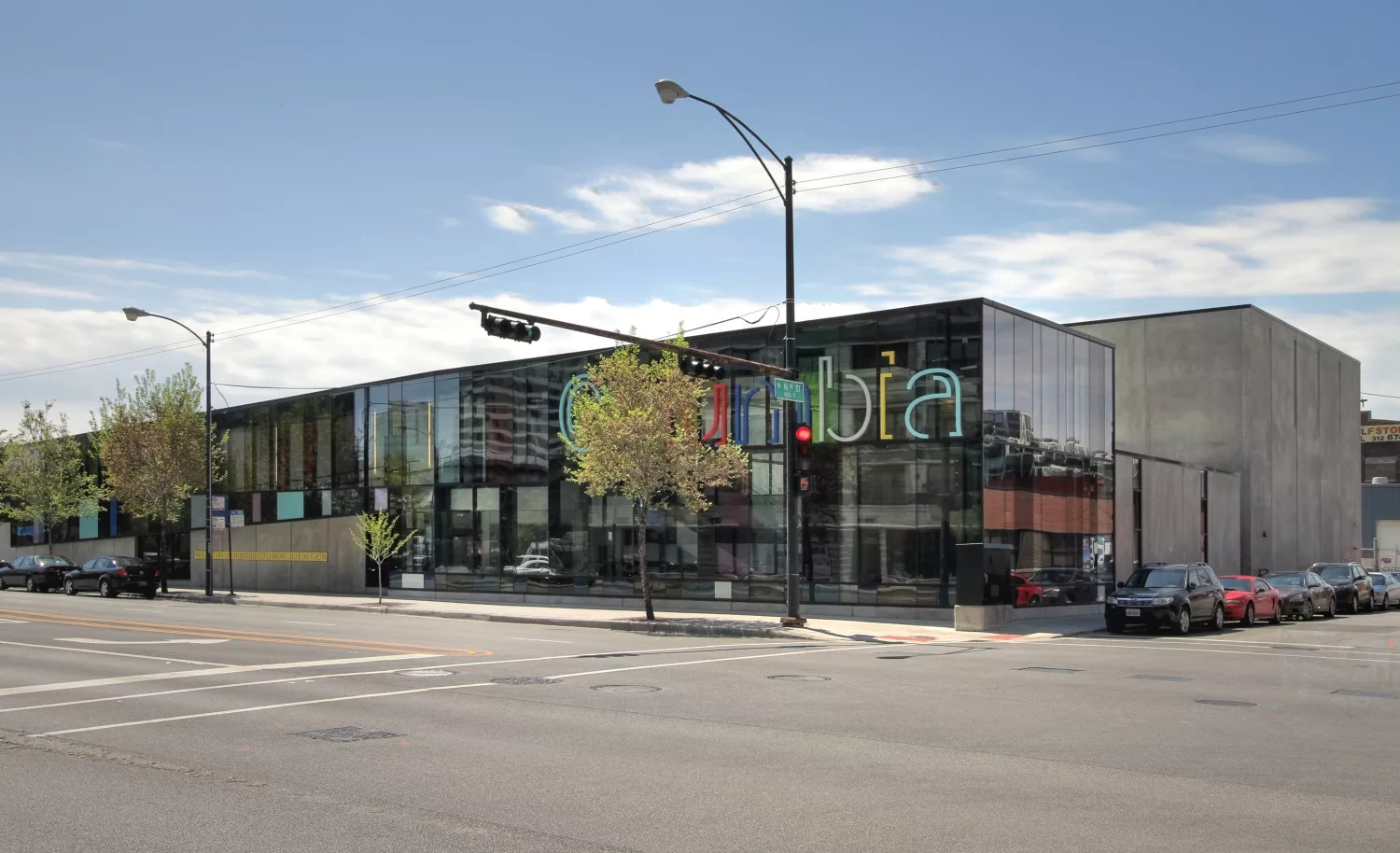 Exterior daylight view of the glass facade of the Columbia College Media Production Center with multi-colored signage and surrounded by parked vehicles