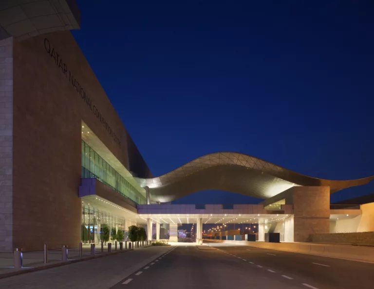 Exterior twilight view of the curved canopy spanning the roadway and pedestrian bridge at the Qatar National Convention Center
