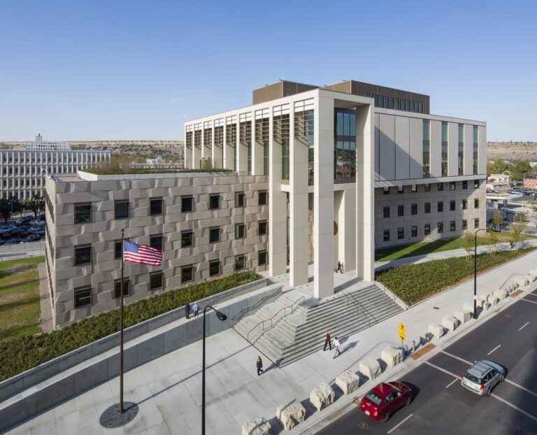 Aerial daylight view of the six-story James F. Battin United States Courthouse featuring rooftop photovoltaic panels, steps leading to the entrance, sidewalk landscaping, and stone bollards adjacent to a two-lane road