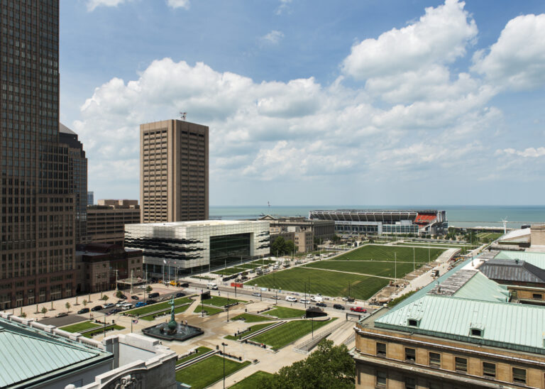 Aerial daylight view of the Cleveland Convention Center and Global Center for Health Innovation with an expansive rooftop lawn, public plaza, and fountain surrounded by high-rise buildings, a football stadium, and Lake Erie in the distance