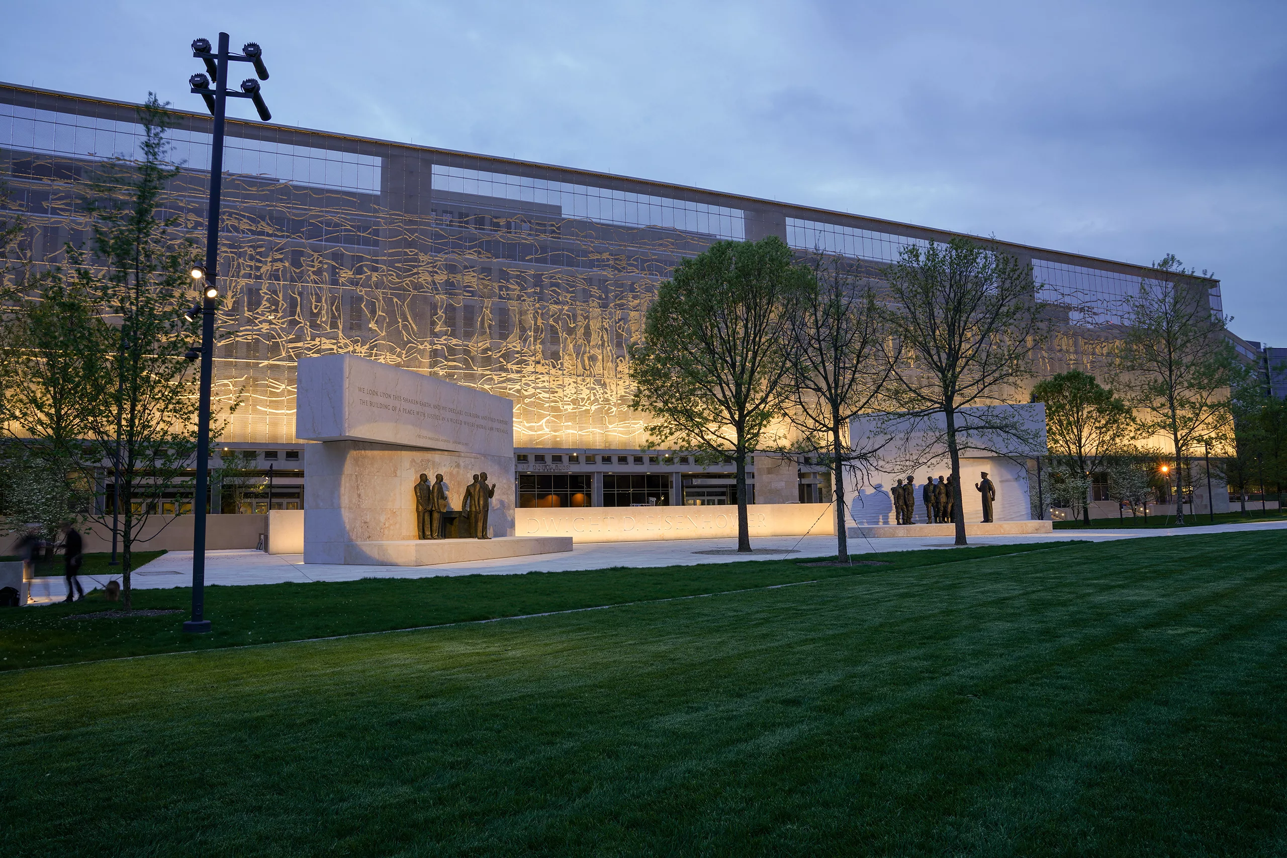 Exterior twilight view of the 80-foot-tall, stainless steel Dwight D. Eisenhower National Memorial featuring dual stone bas-reliefs with engraved quotations and bronze figures, as well as a vast lawn and interspersed trees