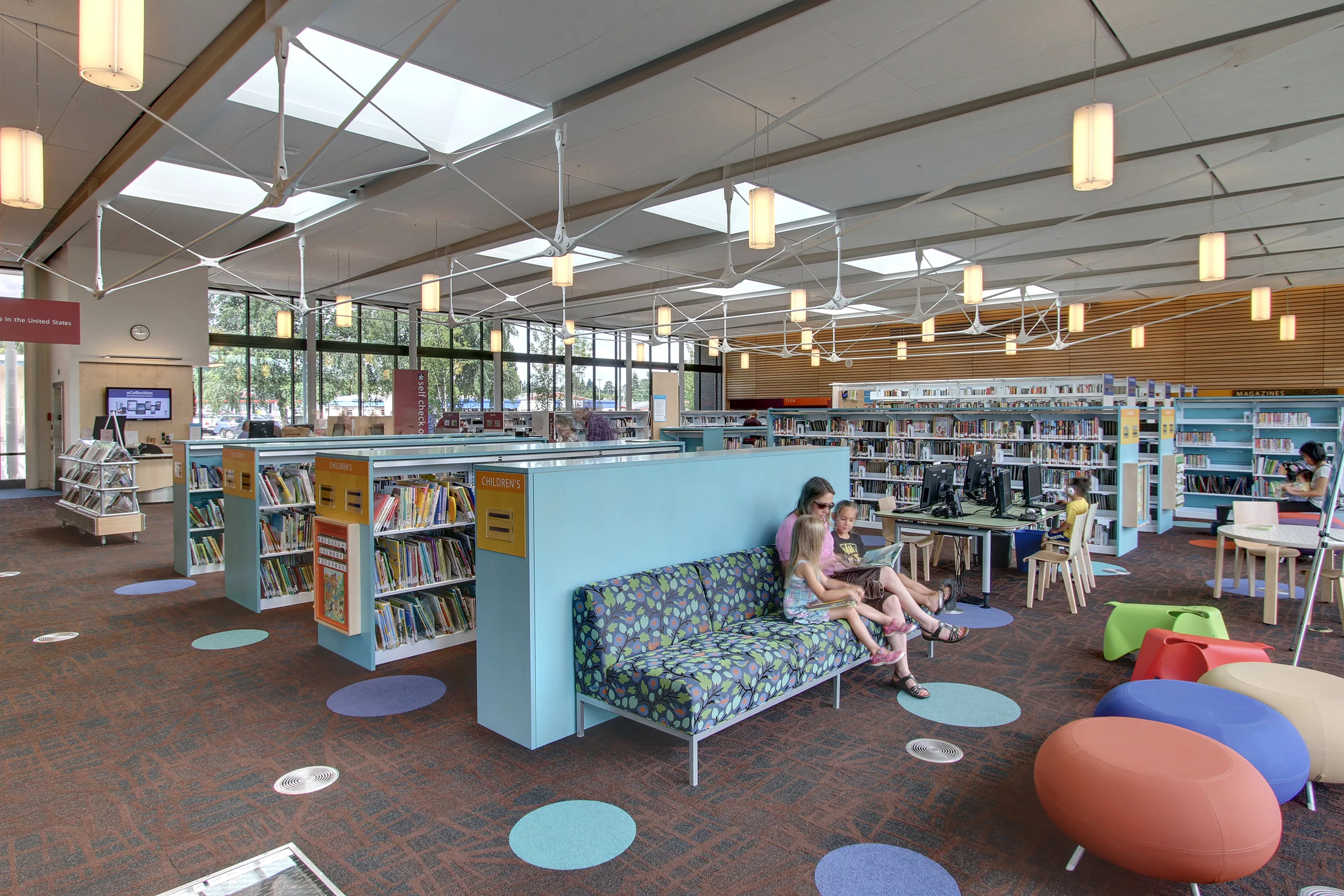 Interior daylight view of the children's reading room at the Kenmore Library featuring bookcases, sofas, children and adults reading, computer stations, and skylights