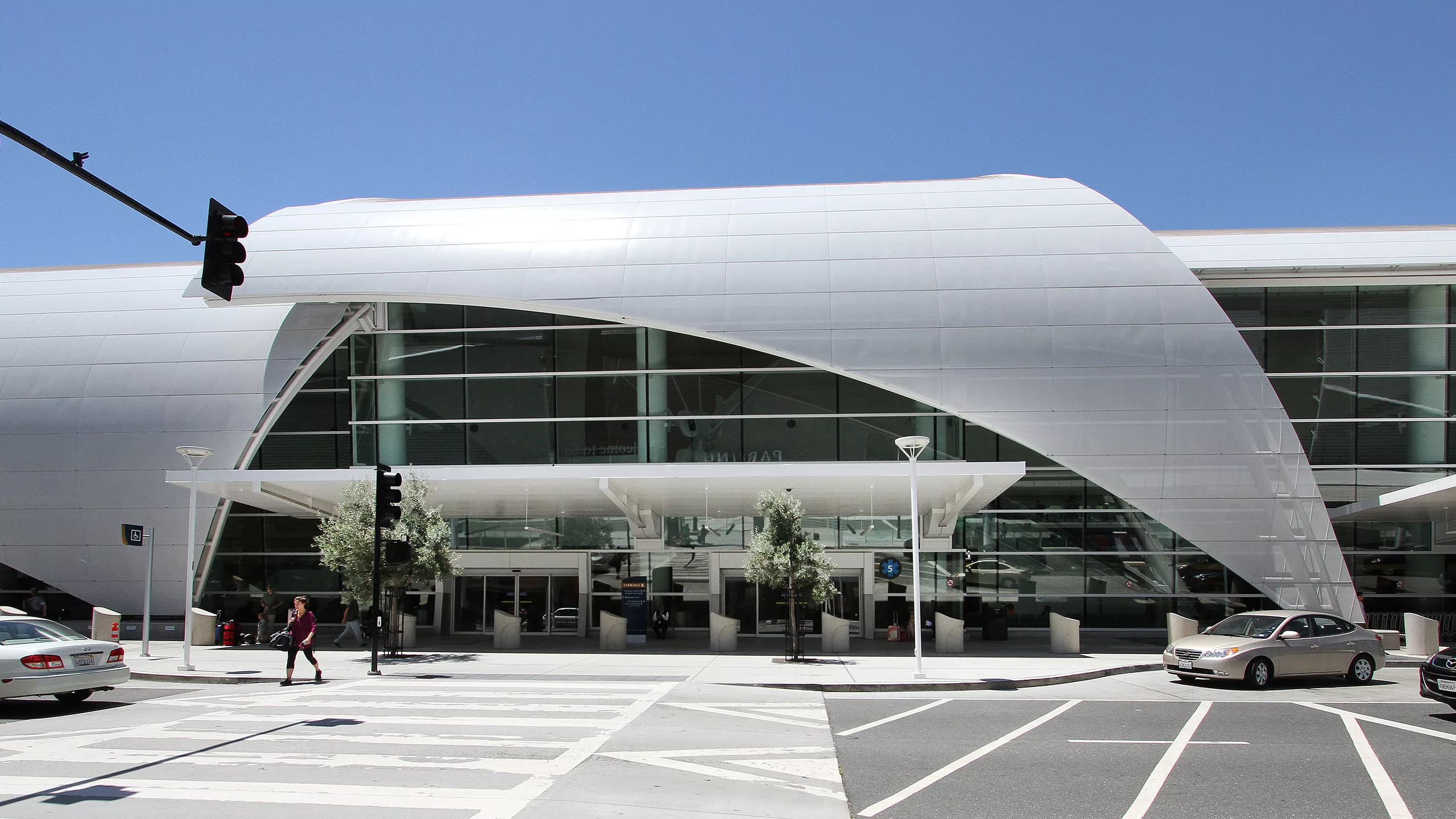 Exterior view of Mineta International Airport's Terminal B in daylight featuring a glass facade partially wrapped in curved metal and several vehicles parked curbside