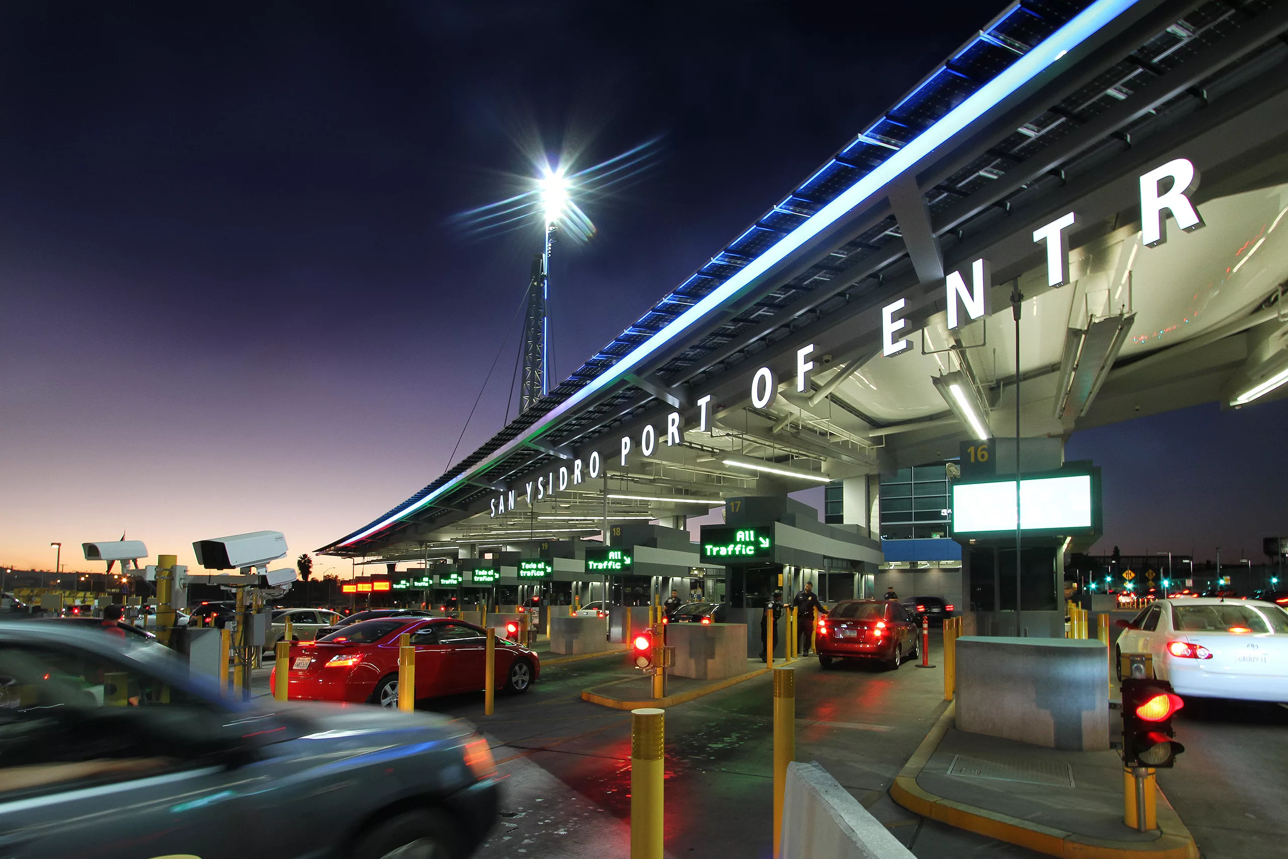 Exterior twilight view of the vehicles lined up in lanes of the San Ysidro U.S. Land Port of Entry with illuminated signage, security cameras, and booths for officers