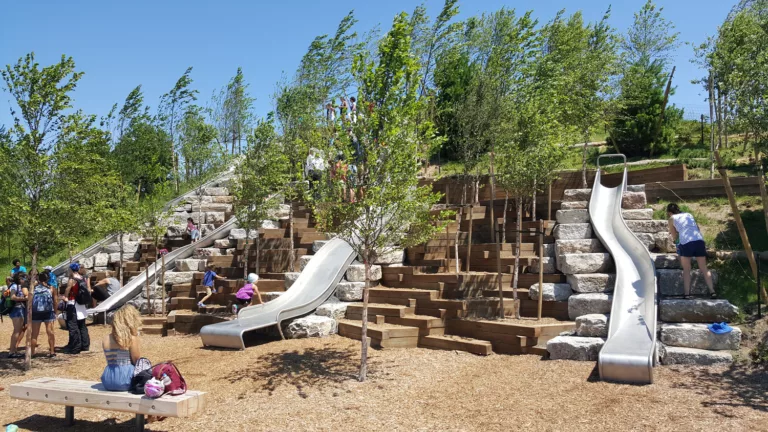 Daylight view of Governors Island Park’s outdoor playground featuring three slides, trees, climbing steps, and a bench, all populated by adults and children