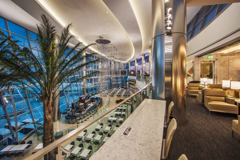 Interior evening image of the downstairs dining hall and an upstairs lounge with an indoor palm tree and ceiling-mounted, cube-shaped public art at San Diego International Airport Terminal 2 West