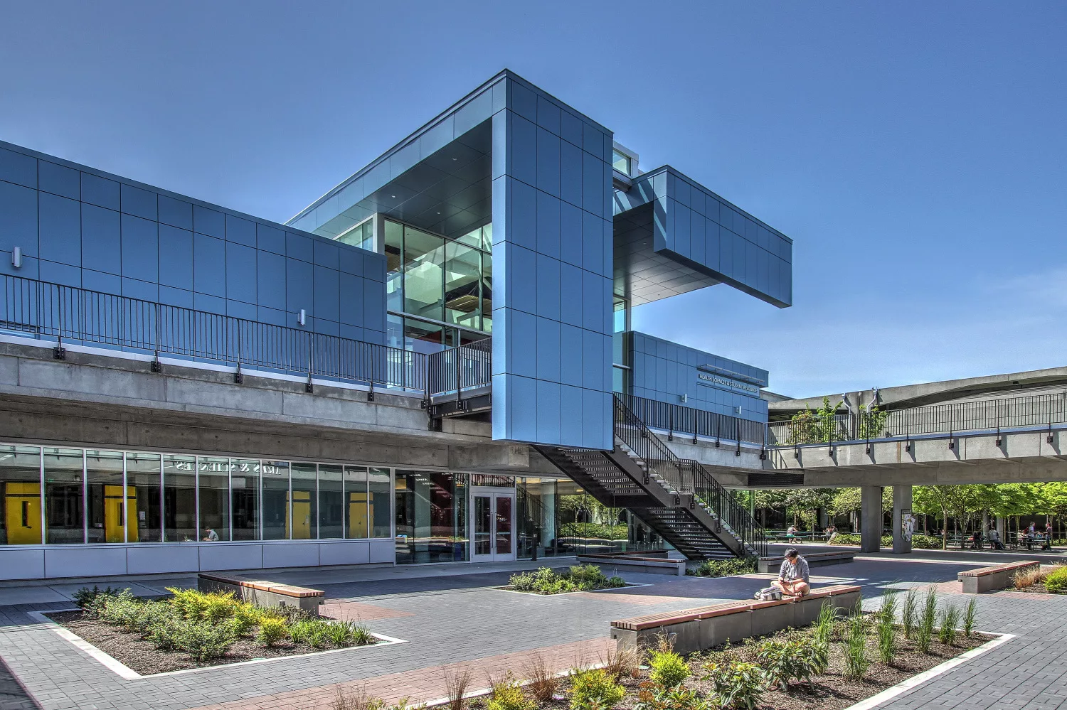 Exterior daylight view of North Seattle College's Health Sciences and Student Resources Building featuring an outdoor plaza with landscaping and benches, a staircase, and elevated walkways for visitors
