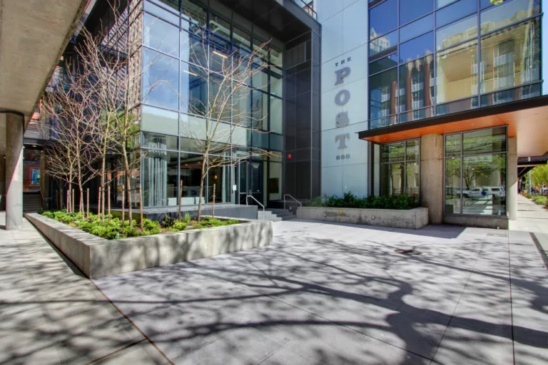 Exterior daylight view of the 16-story Post Apartments' plaza entry with building signage and perimeter landscaping such as trees and small plants