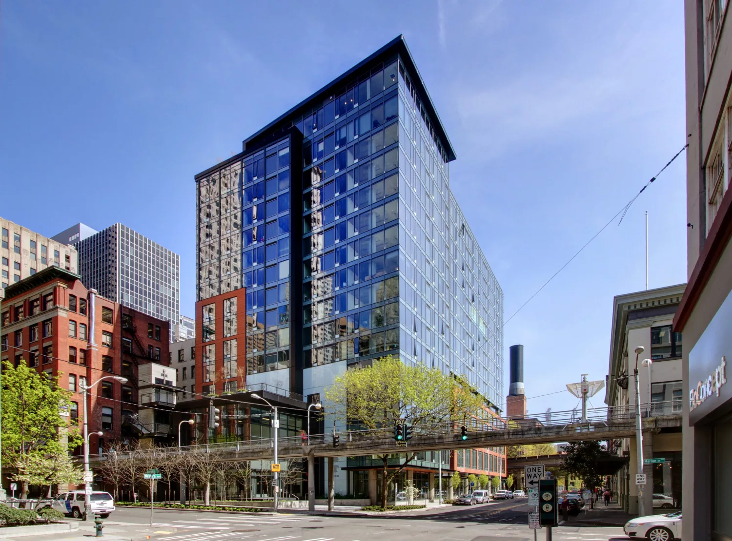 Exterior daylight view of the 16-story Post Apartments' glass facade with an elevated pedestrian bridge in the foreground and surrounding high-rise buildings