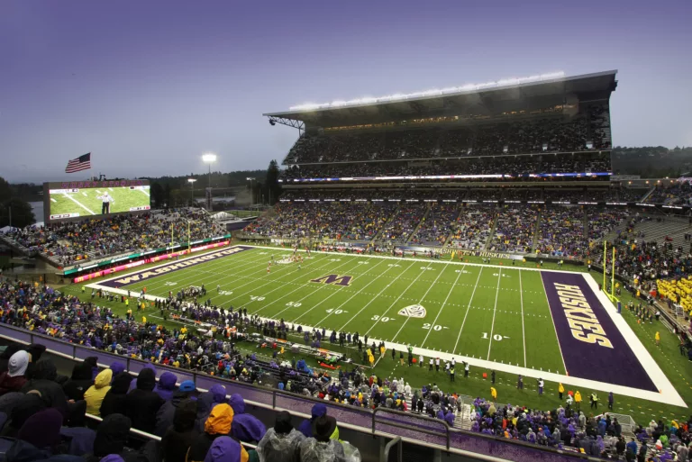 Exterior twilight view of the University of Washington’s open-air Husky Stadium with players on the football field and fans in the multi-level stands and seating bowl