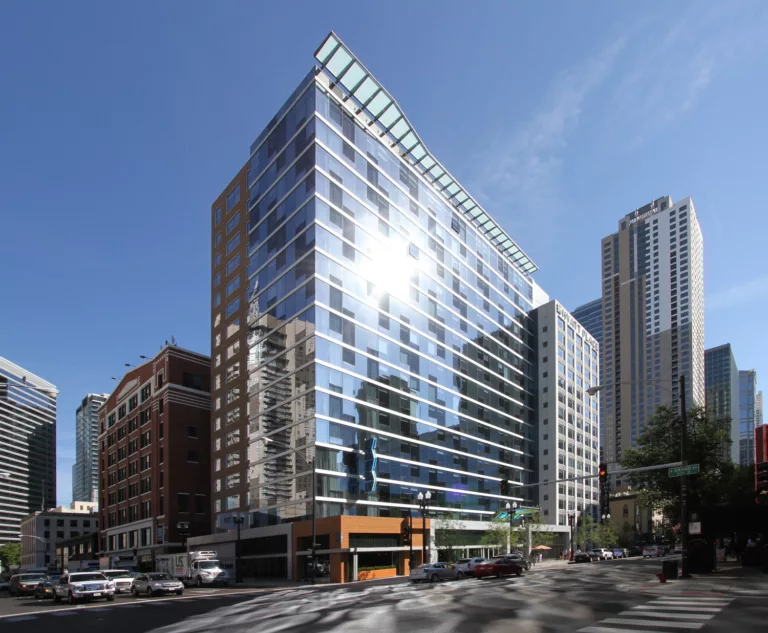 Daylight view looking across the street at the steel-and-glass facade of the 19-story Aloft Hotel at the corner of North Clark Street and West Grand Avenue with surrounding high-rise buildings and vehicle traffic