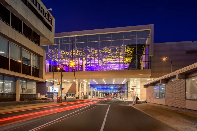Evening exterior view of the Winnipeg Convention Centre Expansion with the purple-and-yellow-illuminated City Room spanning York Street and its pedestrians and vehicles