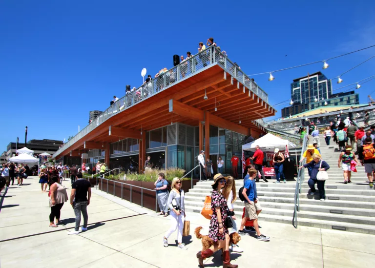 Exterior daylight view of the Pike Place MarketFront Entrance’s public gathering space with visitors walking up a staircase, looking out from a rooftop observation platform, and indoor dining area