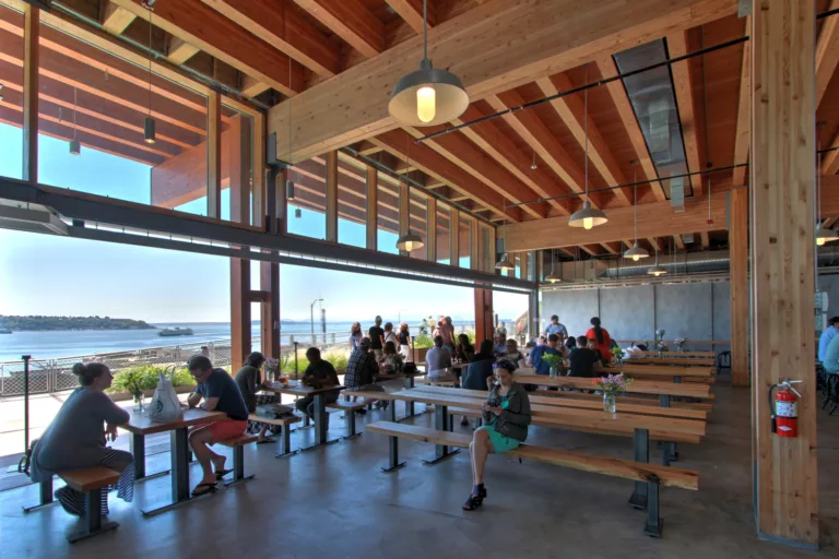 Interior daylight view of the Pike Place MarketFront Entrance’s covered dining area with people gathered at tables, overhead lighting, a wood-beam ceiling, and a ferry on Elliott Bay in the distance