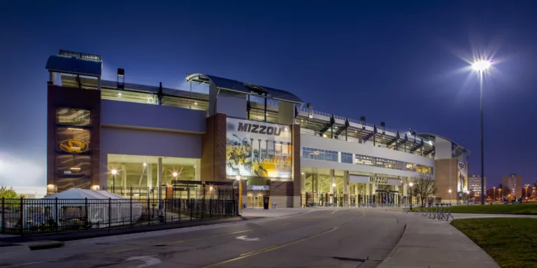 Exterior twilight view of the entrance to the University of Missouri's Memorial Stadium with two-story signage, roadway frontage, bike racks, and lamp posts