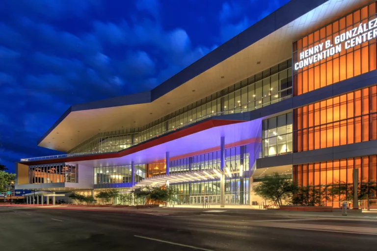 Exterior twilight view of the Henry B. González Convention Center Expansion with a soaring glass-and-steel facade, landscaped trees in the entry plaza, and a roadway