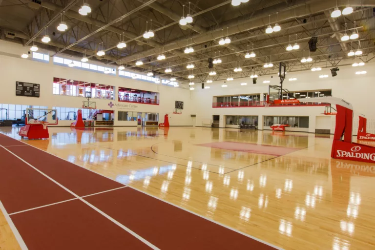 Interior view of several basketball courts at the Chicago Bulls Practice Facility with overhead lights fixed to an exposed steel-frame ceiling, wall-mounted scoreboards, and a second-floor exercise room
