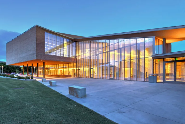 Exterior twilight view of the Greenhill School Marshall Family Performing Arts Center's illuminated and glass-enclosed lobby surrounded by a plaza and central lawn