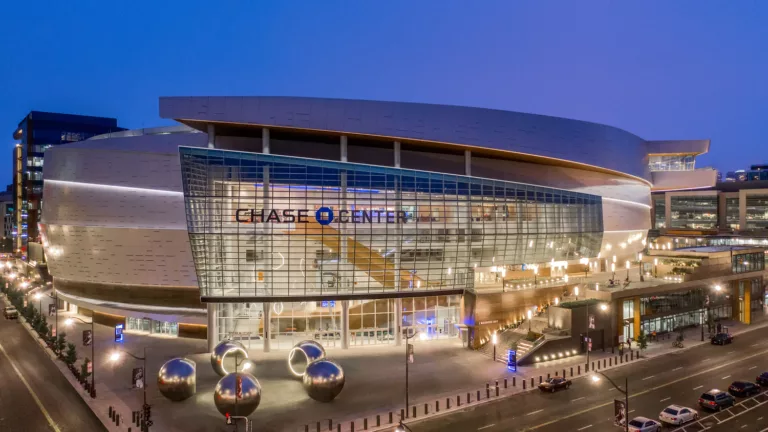 Exterior twilight view of Chase Center’s floor-to-ceiling window facade illuminated within and featuring a plaza with public art and a staircase