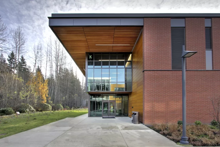 Exterior daylight view of the entrance to Peninsula College's Allied Health and Early Childhood Education Building with a pedestrian walkway, lamp post, and surrounding natural landscape