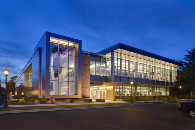 Exterior twilight view of the University of Portland's two-story Beauchamp Recreation and Wellness Center illuminated from within to reveal a climbing wall and exercise equipment inside and boulder-shaped benches, landscaping, and a parking lot outside