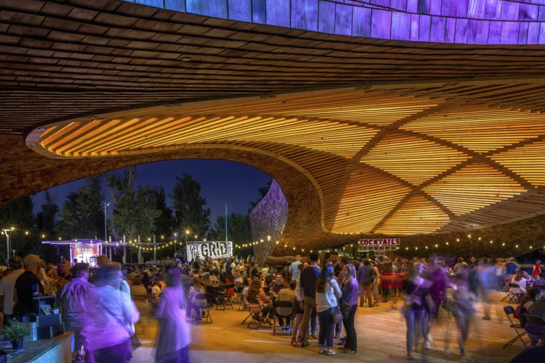 Exterior evening view of a large crowd gathered beneath The Barn's illuminated timber pavilion