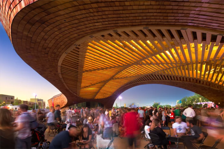 Exterior twilight view of a large crowd gathered beneath The Barn's illuminated timber pavilion