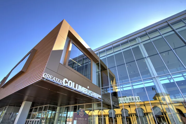 Exterior daylight view looking up at the entrance to the Columbus Convention Center with a soaring glass facade, prominent signage, and a historical marker