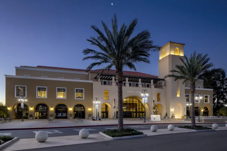 Exterior twilight view of the entrance to the California Baptist University Arena featuring a mounted lancer bronze sculpture, two palm trees, five round bollards, and assorted tables and chairs