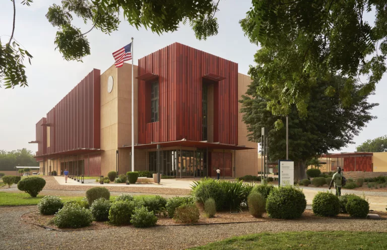 Exterior daylight view of the landscaped entrance to the U.S. embassy in Niamey with signage, a flag pole, and three visitors entering and leaving the building