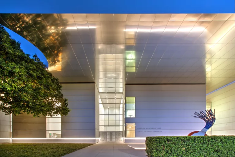 Exterior twilight view of the illuminated steel-and-glass entrance to the Norton Museum of Art Expansion, with landscaping and outdoor public art