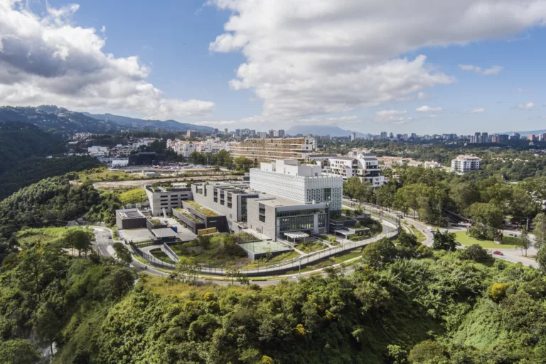 Exterior of the U.S. Embassy in Guatemala City surrounded by terraced trees and lush landscaping, a sloping driveway bordered by black fencing