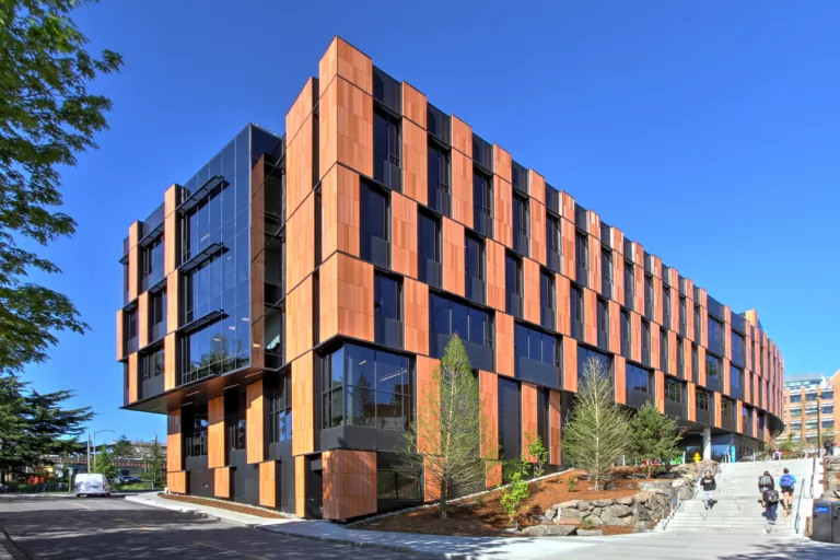 Exterior daylight view of the University of Washington's five-story Bill & Melinda Gates Center for Computer Science & Engineering with a glass-and-wood facade and surrounding roadway, staircase, and hillside landscaping