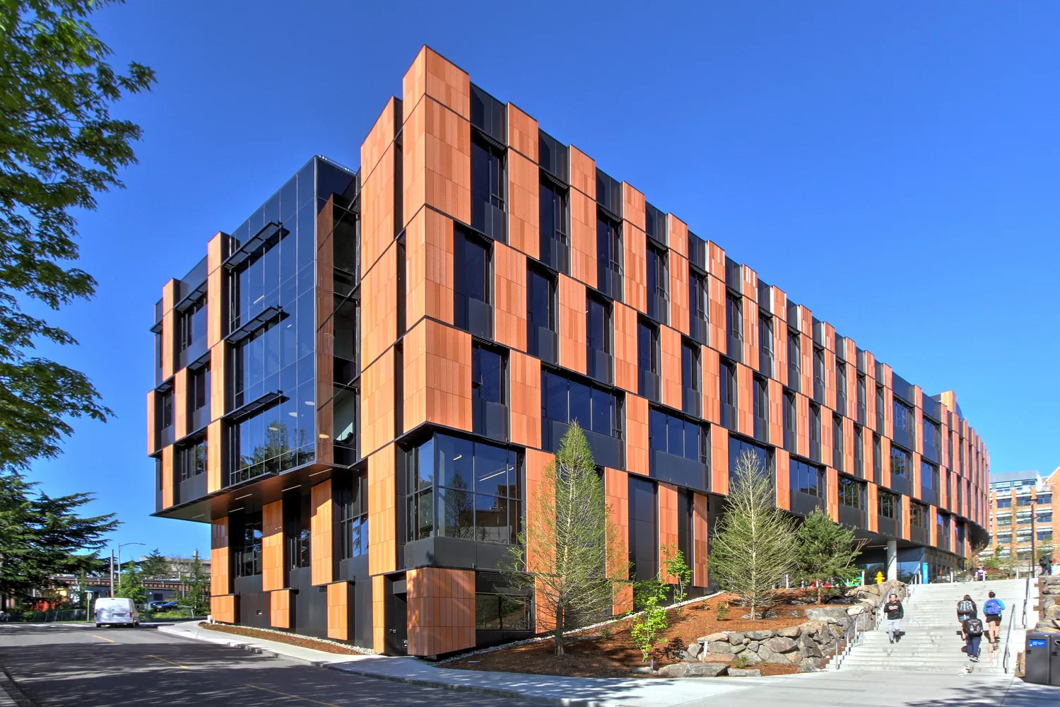 Exterior daylight view of the University of Washington's five-story Bill & Melinda Gates Center for Computer Science & Engineering with a glass-and-wood facade and surrounding roadway, staircase, and hillside landscaping