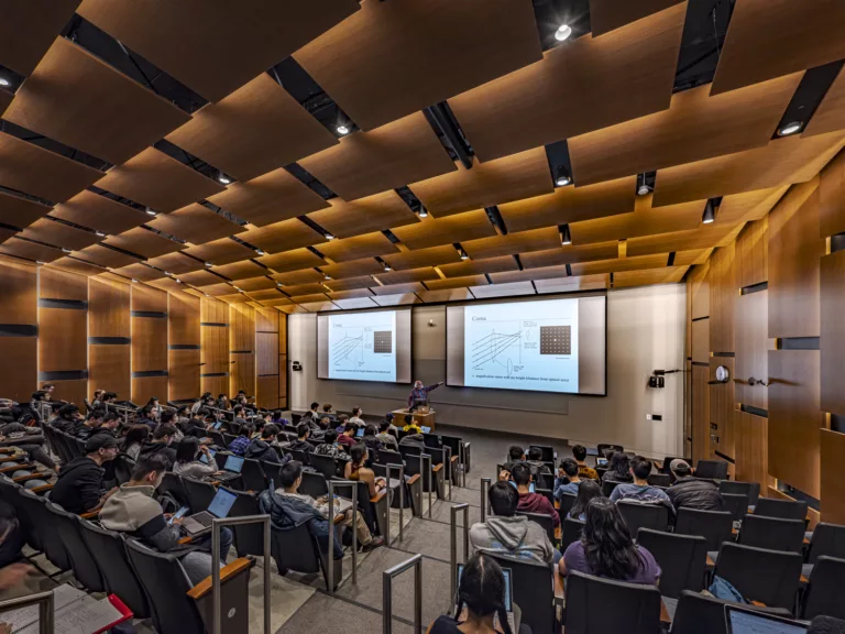 Interior view of a student-filled classroom at the University of Washington's five-story Bill & Melinda Gates Center for Computer Science & Engineering featuring raked seating, two display screens, and wood-paneled overhead lighting