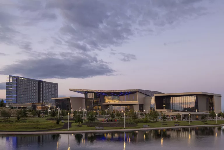 Exterior twilight view of the Oklahoma City Convention Center and its angular rooflines, with a public park, landscaping, and waterfront trail with benches and street lamps