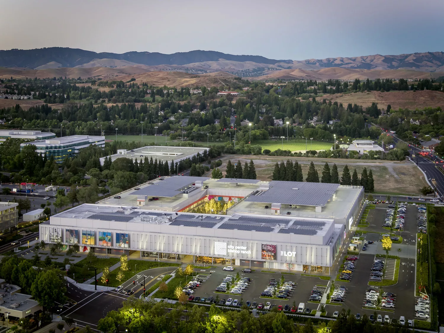 Aerial twilight view of the two-story City Center Bishop Ranch and its central outdoor piazza with surrounding surface parking lot and distant hills