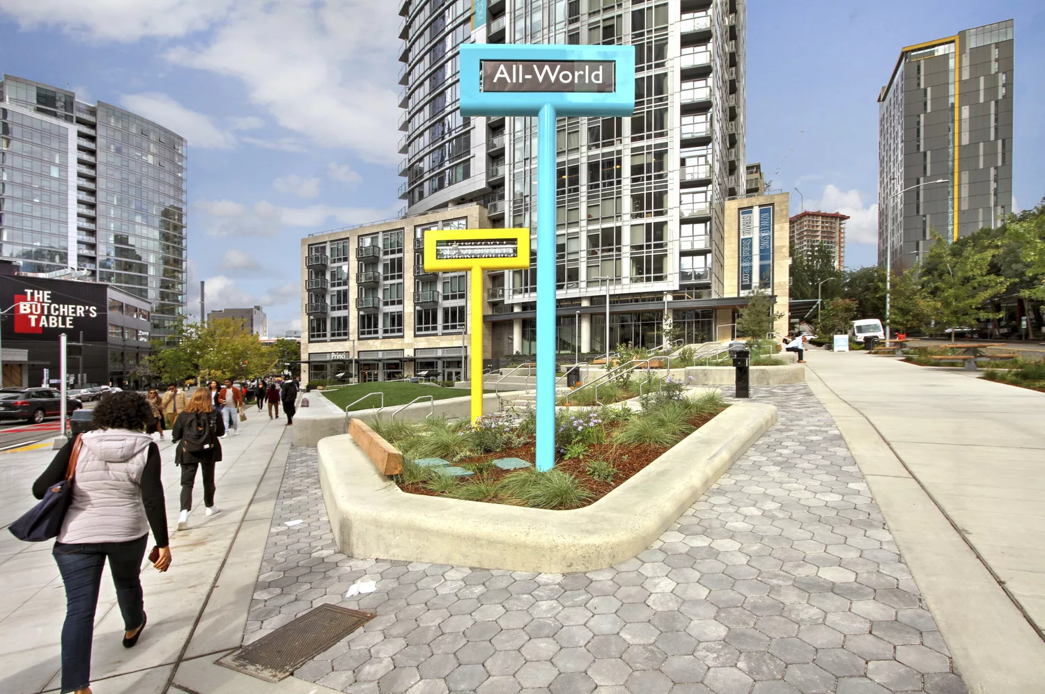 A daylight view of Urban Triangle Park’s colorful signage, steps, seating areas, landscaping, and surrounding high-rise office and residential buildings